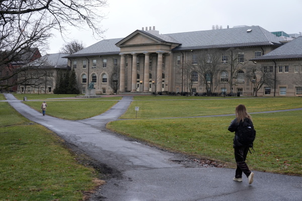 Two women wearing backpacks walk on a paved pathway on the campus of Cornell University in Ithaca, N.Y., in 2024. The pathway cuts through an open area with green grass. In the background is an old-style, three-story building with four pillars.