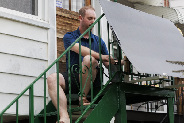 Sitting outdoors on his back steps, which are metal and painted green, Craig Keenan installs a plug-in solar panel in front of the railing at the top.