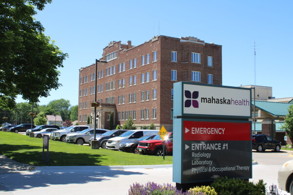 A roughly four-story, old-looking brick building stands in the background with a row of cars parked in front of it. In the foreground is a sign displaying the name Mahaska Health and arrows directing people to the emergency room and Entrance 1.