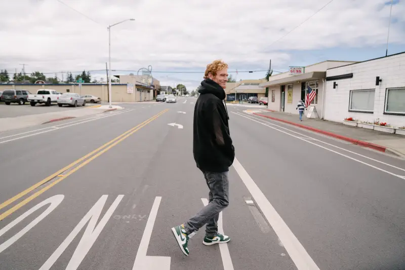 Cody Coughenhour crosses a street in Port Angeles, Wash. He's wearing a black hoodie and a cloudy sky is visible in the distance.