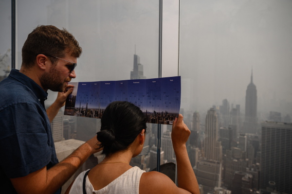Visitors hold a map showing city landmarks in a clear day as they stand before the Manhattan city skyline during heavy smog brought by wildfire smoke from Canada. Climate change is making wildfires likely to burn more intensely; their smoke is damaging air quality in the U.S. West as well as in parts of the country that have not historically dealt with smoke, like New York City.