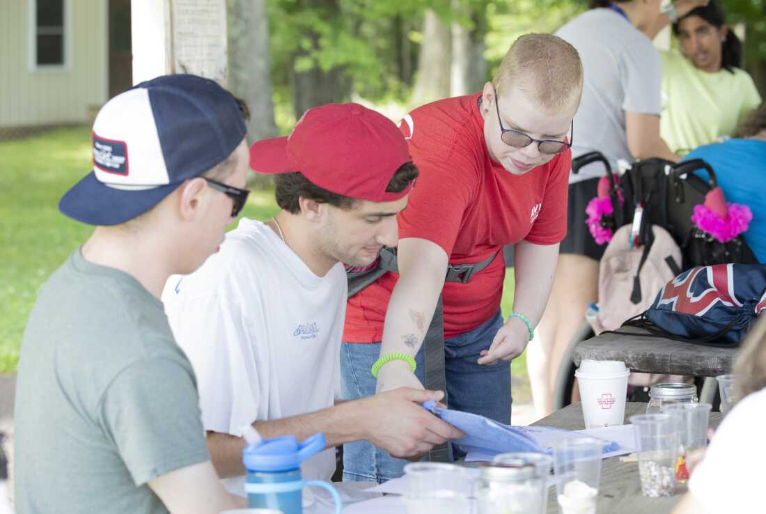 Brianna Engleman stands outdoors next to a wooden picnic table, at which are seated two adolescent male campers.