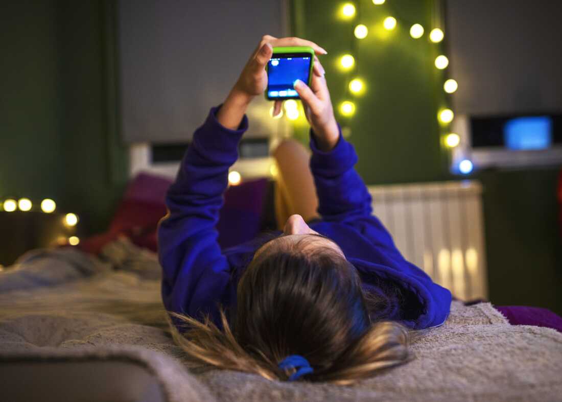 A girl wearing a long-sleeved blue shirt lies on her bed, holding her phone. Her bedroom is decorated with strings of white lights.