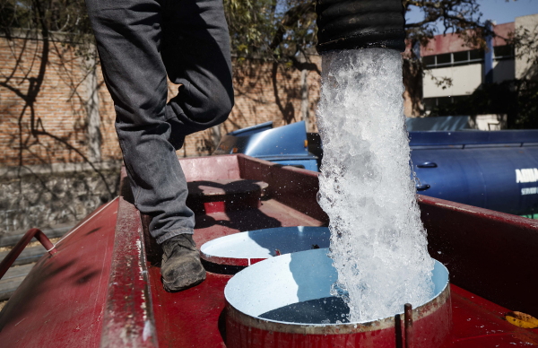 In this photo, a worker with Mexico's National Water Commission fills a water truck with drinking water to be distributed in Mexico City in January 2024. The man, photographed from the waist down and wearing dark pants and boots, is standing on a water truck while water pours into a hole on the top.