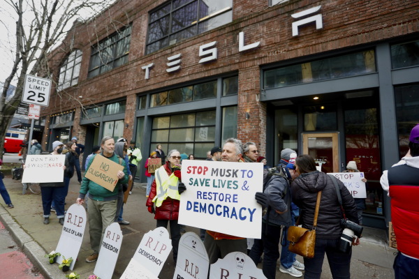 People protesting Musk's actions in the Trump administration stand in front of signs shaped like tombstones with phrases such as "RIP Democracy." One man holds a sign that says "Stop Musk To Save Lives & Restore Democracy." They are protesting outside a Tesla showroom in Seattle on Feb. 13.