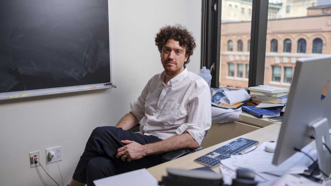 Tristan Buckmaster, mathematician at New York University, poses for a portrait in his office in Manhattan. He is wearing a white button down shirt and dark slacks. Behind him is a blackboard and a window. In front of him is a desk with a computer, keyboard and some papers.