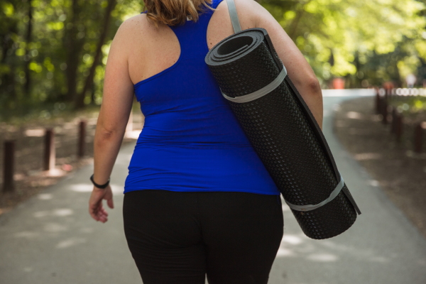 A woman is shown from the back. She's wearing workout clothes and is carrying a rolled-up exercise mat.