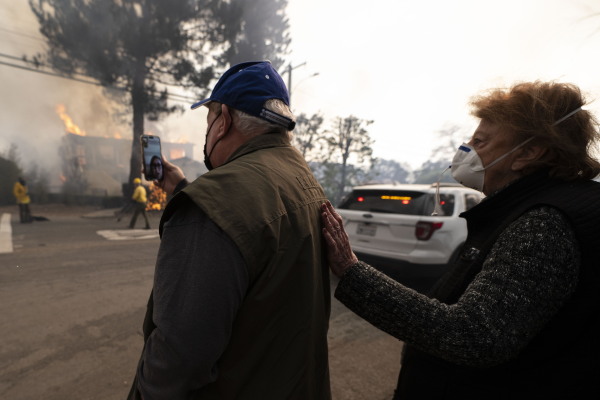 In this photo, an older man holds a smartphone during a video call with a loved one. A woman wearing a mask stands behind him. In the background, a structure is on fire, with flames shooting from the top of it.