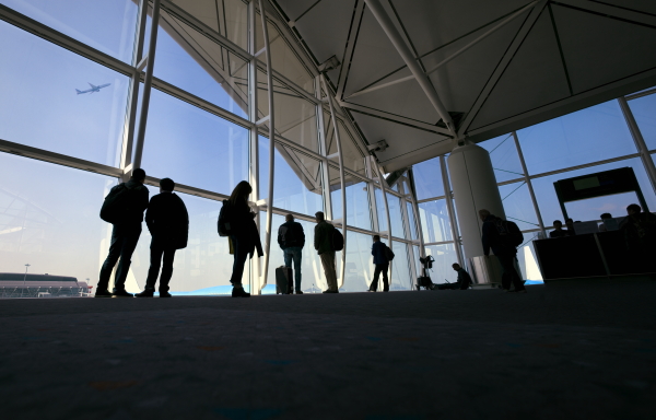 Low angle photograph of silhouettes of people waiting in line at the airport gate. The space has large windows and an airplane flies high in the distance.