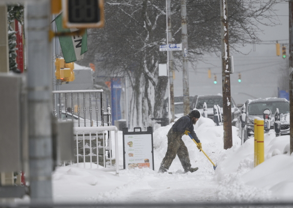 A man shovels snow off a sidewalk. Tall heaps of snow line the curb along the street.