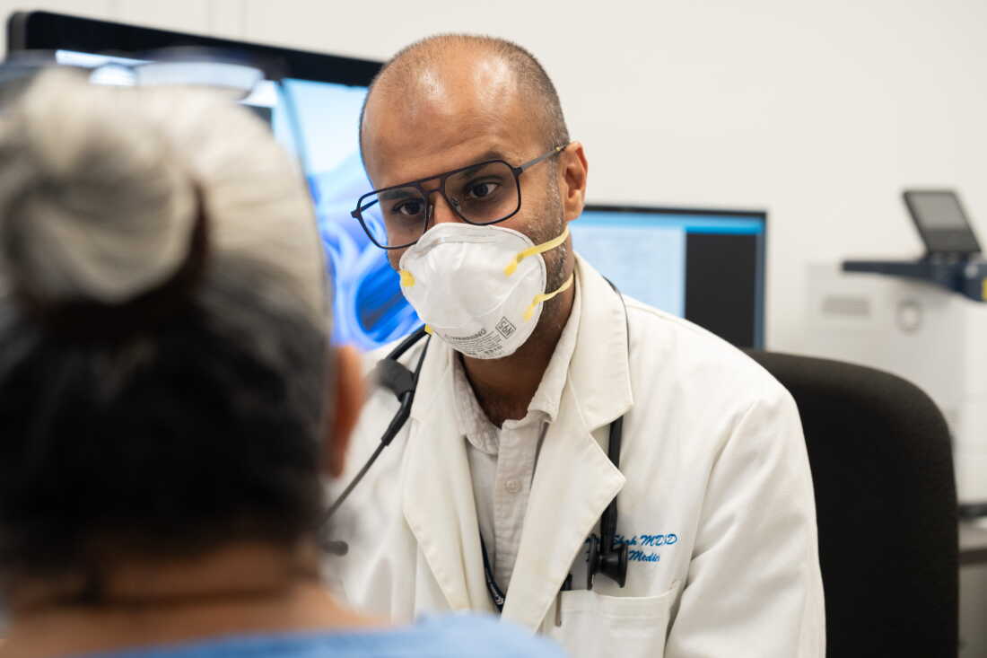 Physician Naman Shah wears a mask and a white coat at Monrovia Health Center in Monrovia, California. A stethoscope is draped over the back of his neck.