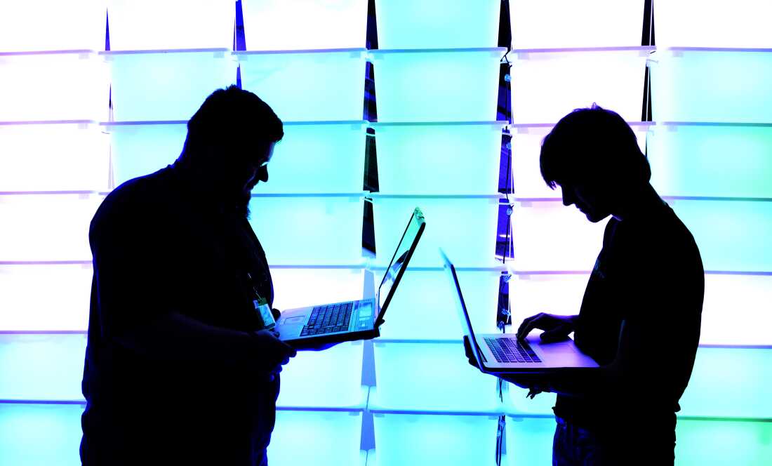 Two men stand silhouetted against an illuminated blue and purple background. They are facing each other, but looking down at laptops they are holding in their hands. 