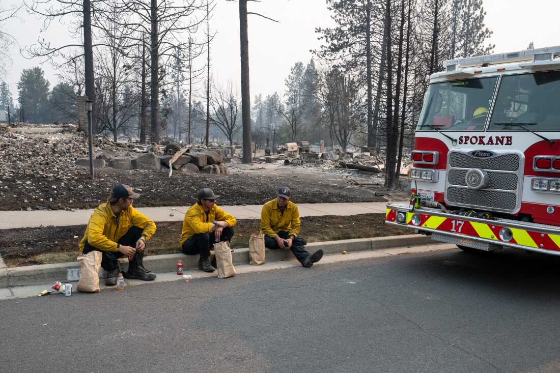 Three members of the Spokane Fire Department sit on a curb in front of burned out residential homes in the Balboa neighborhood of Spokane, Washington, on August 2, 2026. Fast-moving fires left a trail of destruction in the northwestern US city of Spokane on Sunday, with officials reporting hundreds of structures reduced to cinders. Thousands of people were ordered to evacuate their homes Saturday and overnight into Sunday, as three separate fires broke out in and around the city in the east of Washington state.