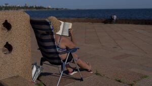 A woman wearing a hat and bathing suit while sitting in a folding lawn chair reads a book and sunbathes near a body of water.