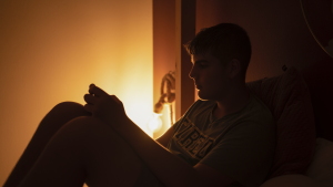A teen boy is shown sitting up in his bed, looking at his phone. The room is dark except for dim lamplight.