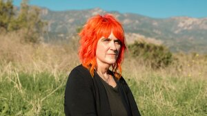 Micky Small53is a screenwriter and is one of hundreds of millions of people who regularly use AI chatbots. She has bright orangeshoulder length hair and is seated with a background of mountains and brush behind her.