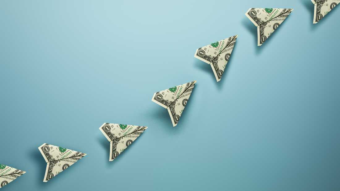 A photograph of seven one dollar bills folded into the shape of a paper airplanes on a blue background. The airplanes are lined up from the bottom left of the frame to the top right creating a trail.