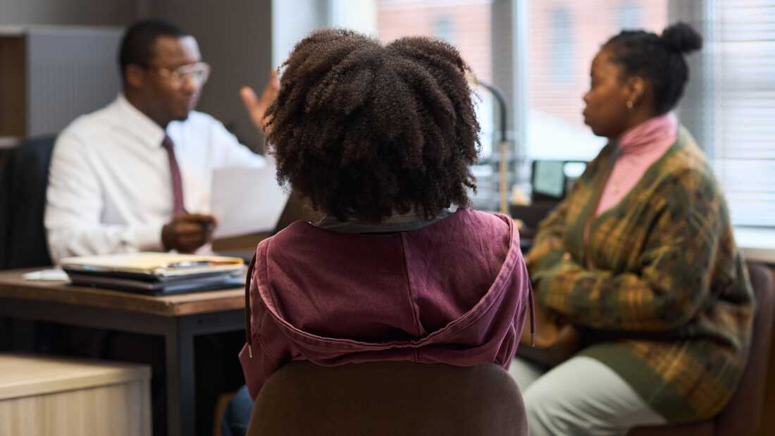 Black man appearing middle aged sitting at desk talking to Black woman and Black child in office setting, principal discussing school matters with parent and student
