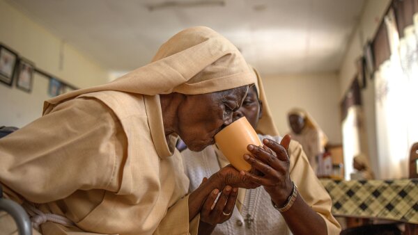 One nun assists an elderly nun as she takes tea. They are wearing yellow-toned apparel.