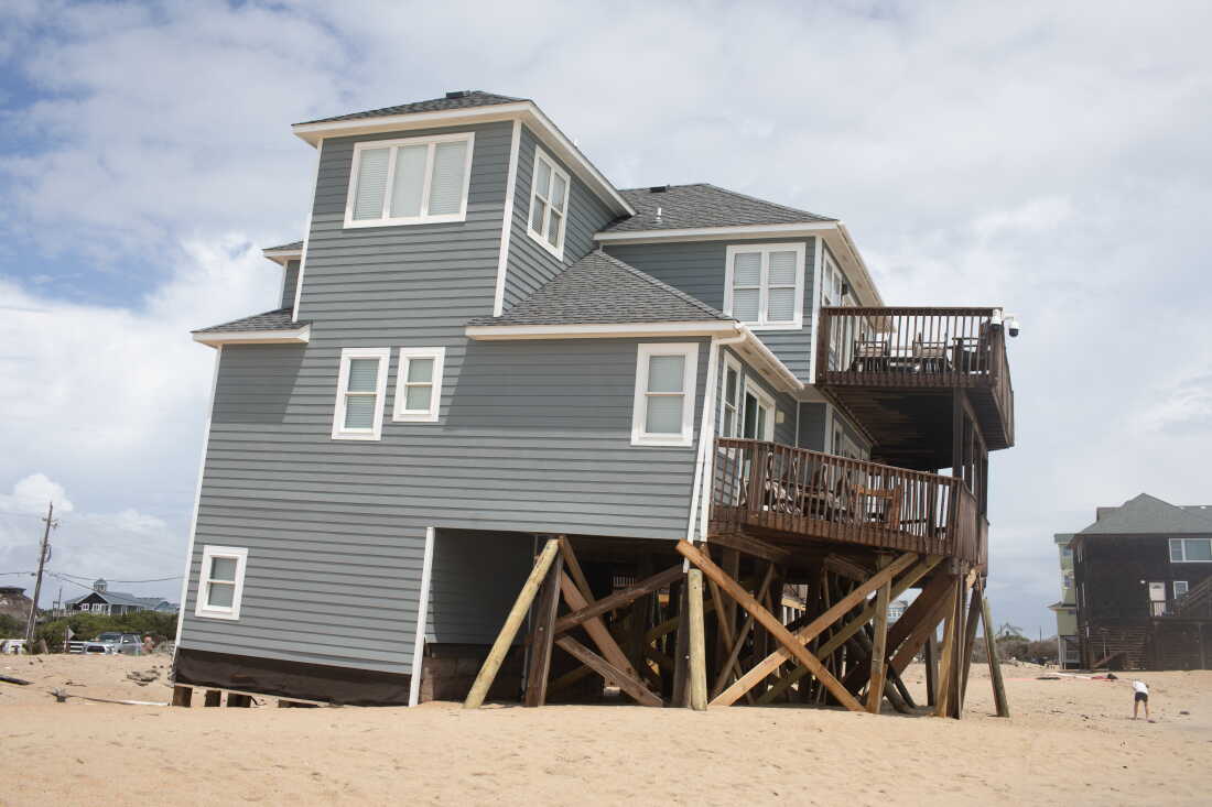 A three-level house with gray siding is tilted askew as one side of it, supported by wood stilts underneath, sinks into the sand.