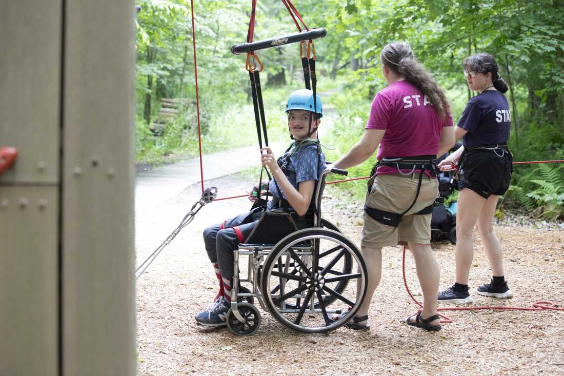 Lucas Hardy is strapped into a hoist while seated in a wheelchair near the rock wall at Camp Cranium. He's wearing a light blue helmet, and two staff members are assisting with the hoist's rope.