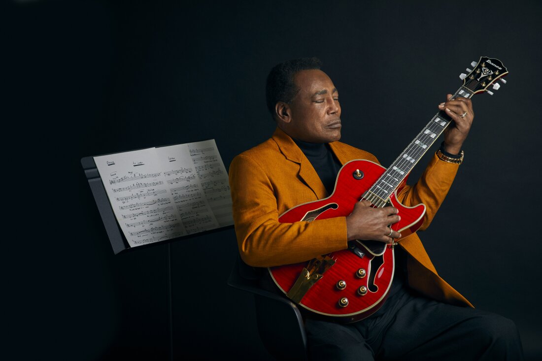 George Benson, dressed in a dark orange blazer jacket, sits holding his guitar in front of a music staned