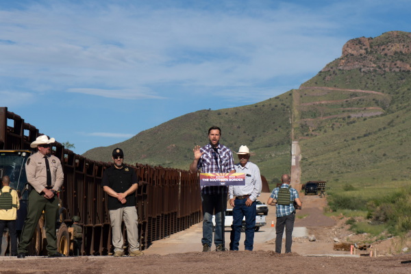 In this photo, Sen. JD Vance of Ohio, the Republican vice presidential nominee, speaks into a microphone attached to a lectern. He is outdoors at the U.S.-Mexico border in Hereford, Ariz., on Aug. 1, 2024. He's wearing jeans, a plaid shirt and boots. On the left is a border fence, and a tall green hill is in the background. A law enforcement official in a uniform and cowboy hat stands near the fence.
