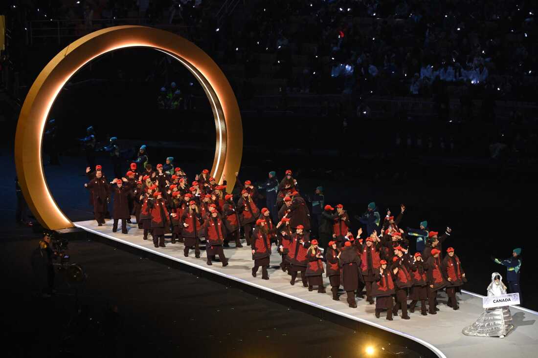 Dozens of Canadian athletes march into the stadium. They are wearing brown jackets with a large red maple leaf, brown pants and red hats.