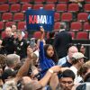 In this photo, supporters attend a Harris-Walz campaign event at Desert Diamond Arena in Glendale, Ariz., on Aug. 9. The photo shows a group of people standing together with rows of arena seats in the background. A girl in the middle of the crowd is holding up a sign that says "KAMALA" in all capital letters.