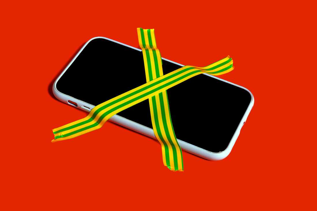This image shows a smartphone with its screen darkened and two strips of yellow-and-green-striped tape arranged in an X shape taping down the phone against a red surface.