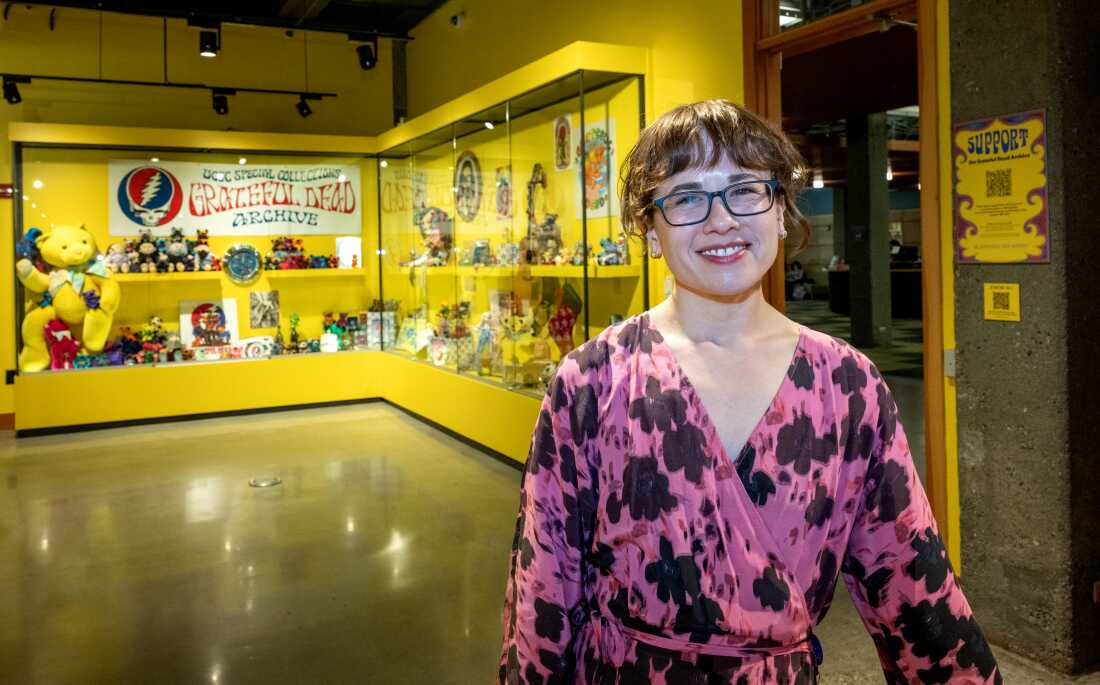 Sam Regal stands in an exhibition space with yellow walls and glass display cases. Grateful Dead memorabilia such as a yellow plush "dancing bear" is in the display cases. Regal is wearing glasses and a pink dress with a dark floral pattern.