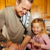 A triptych of photos that shows David Nayfeld making pancakes with his daughter in their kitchen at a wooden island. The first photo shows them putting vanilla extract into a metal mixing bowl. The second photo shows David cutting pancakes on a plate. And the third photo shows his daughter feeding him a bite of pancake.