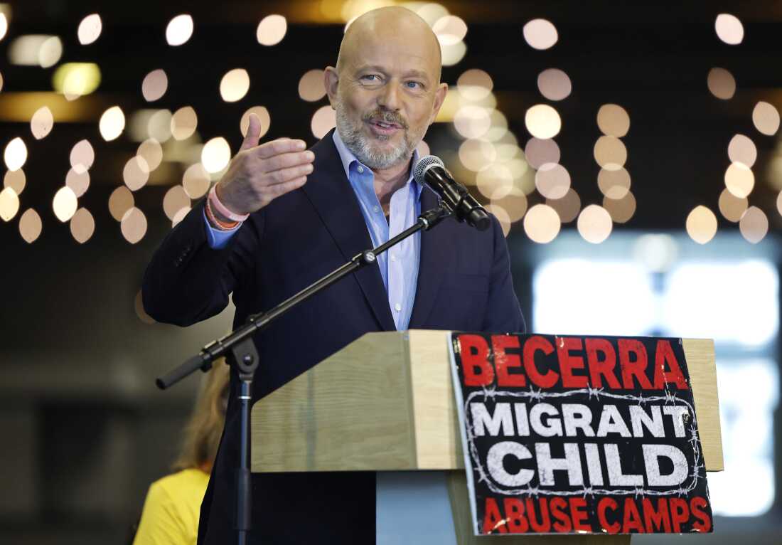 Steve Hilton is shown at a podium with garland lights behind him. A sign on the front of the podium says, "Becerra Migrant Child Abuse Camps."