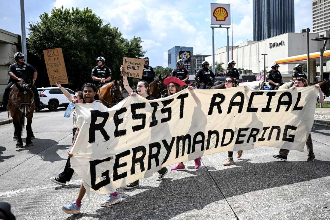 Protesters hold a long, white banner reading "Resist Racial Gerrymandering" in black capital letters as they march in a street during a Labor Day rally in Houston on September 1, 2025. In the street behind the protesters are police officers who are mounted on horses.