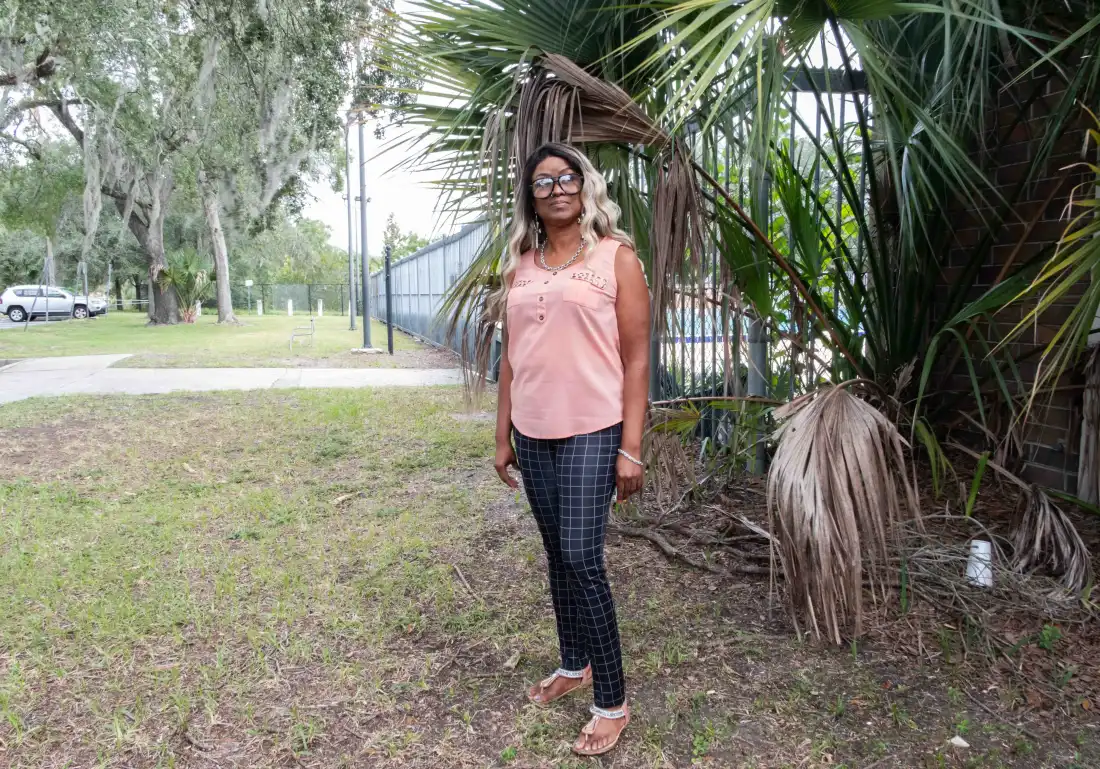 Rachel Dues, a volunteer with Florida Rising, is wearing large glasses, a pink sleeveless blouse and plaid pants. She has black and blonde hair. She is standing next to a fence and a large plant.
