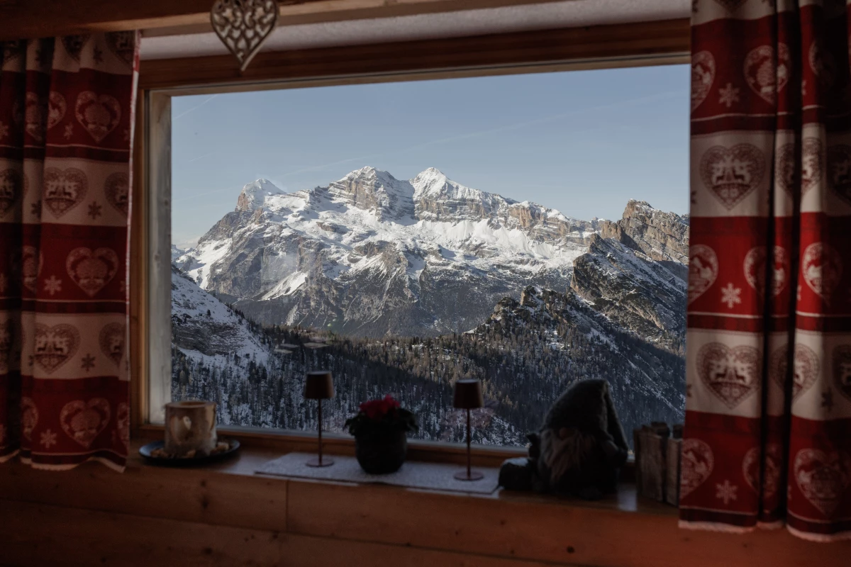 The snow-capped peaks of the Tofane massif are framed through a window of a rifugio, a kind of traditional mountain hut, decorated with typical heart-patterned Ampezzo textiles. These high-altitude lodgings serve as essential rest areas for skiers and hikers, offering a blend of rustic hospitality and panoramic views that define the winter experience in the Dolomites.