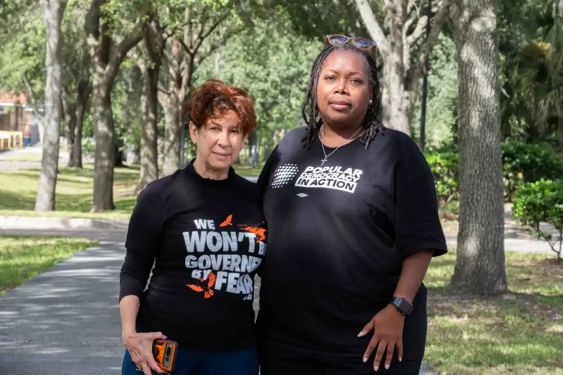 Florida Rising volunteers Maggie Fernandez, 68, (left) and Tia Bell-Williams, 55, stand together in a tree-lined area. Fernandez is wearing a black t-shirt that says "We won't be governed by fear" and Bell-Williams is wearing a black t-shirt that says "Popular democracy in action."