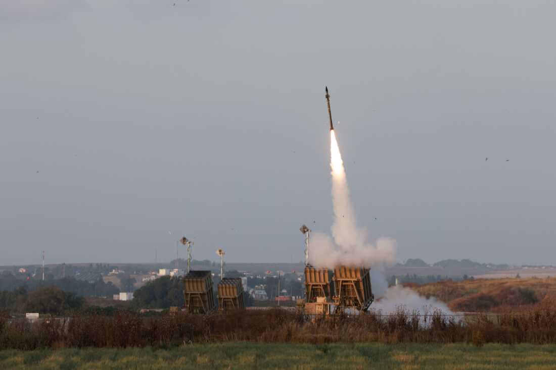 Missile launchers stand in an open grassy area, and one of them launches a missile up into the air against a blue sky. Some buildings appear far away in the background.