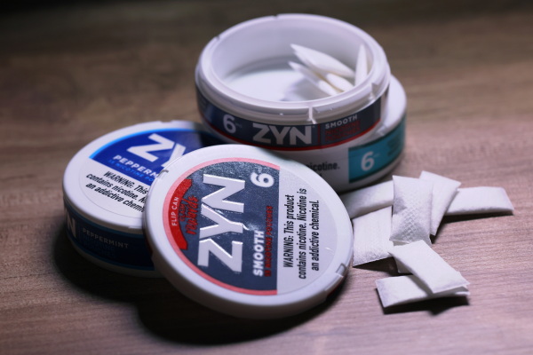 This photo shows two round, puck-shaped containers of Zyn nicotine pouches stacked together. The lid has been removed from the top container, and small white pouches of powdered nicotine are inside.
