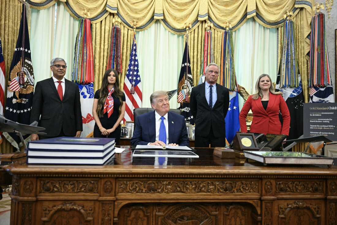 President Donald Trump sits at the Resolute Desk with several people behind him.
