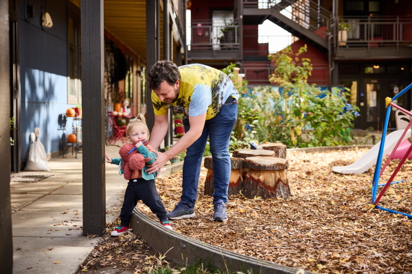 A dad helps his toddler navigate a low playground barrier. The dad is wearing a tie-dye t-shirt; the little girl holds a red stuffed-animal toy.