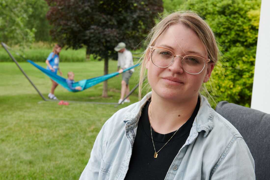 Amber Petersen, ailesinin Iowa City, Iowa'daki evinin arka bahçesinde oturuyor. Sarı saçları geriye toplanmış ve gözlük takıyor. Kocası ve çocukları arkasında hamakta oynuyorlar.