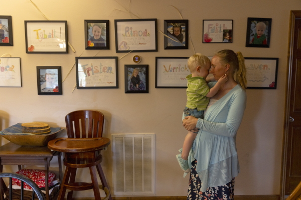 The Algers' youngest child, Sven, who looks to be toddler age, is being held by his mother, and their faces are nearly touching, nose to nose. Framed photos of the family's children hang on the wall behind them, and a wooden high chair is situated against the wall.