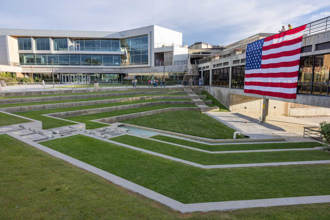 A large U.S. flag in memory of Charlie Kirk hangs on the side of a building on the campus of Utah Valley University in Orem, Utah, on September 19, 2025. The flag faces a terraced, grassy green space.