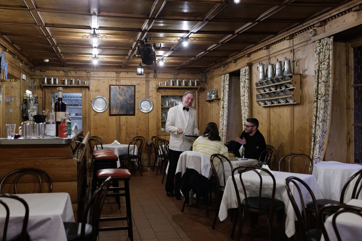 A waiter in a formal white jacket serves patrons inside the wood-paneled Bar Posta at the historic Hotel de la Poste. A cornerstone of Ampezzo social life since the 19th century and a favorite haunt of Ernest Hemingway, the bar represents the 'old-world' heritage of Cortina