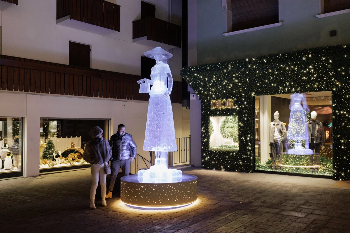 A glowing, life-sized ice sculpture of a woman in an elegant dress and hat stands illuminated at night in a pedestrian area, with the high-end Dior boutique visible in the background. As the town prepares for the Milano Cortina 2026 Winter Olympics, the presence of major global luxury brands underscores the resort's status as an elite international destination