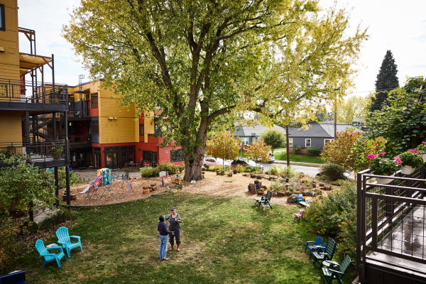 The photo overlooks a large courtyard with a beautiful tree in its center. Two people stand chatting, one has a coffee cup in her hand.