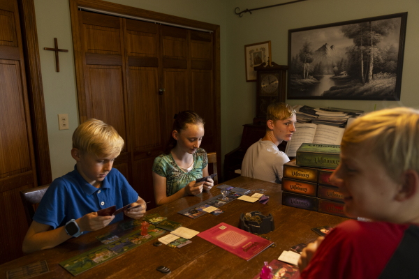 Three of the Algers' older children, Tybalt, Aurora and Dimitry, are seated at a wooden table and are playing a card-based game. The eldest child, Aiden, is in the background and is seated at a piano.