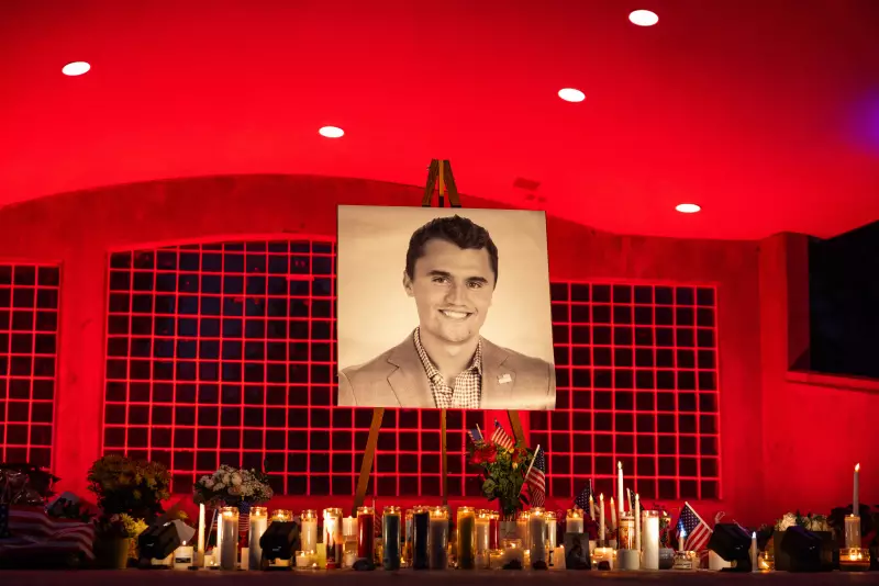 A large black and white photo of Charlie Kirk, 31, is seen at his makeshift memorial at Orem City Center Park in Orem, Utah. The area is lit with red lights and there are dozens of lit candles underneath the photo along with a few American flags and flower bouquets.