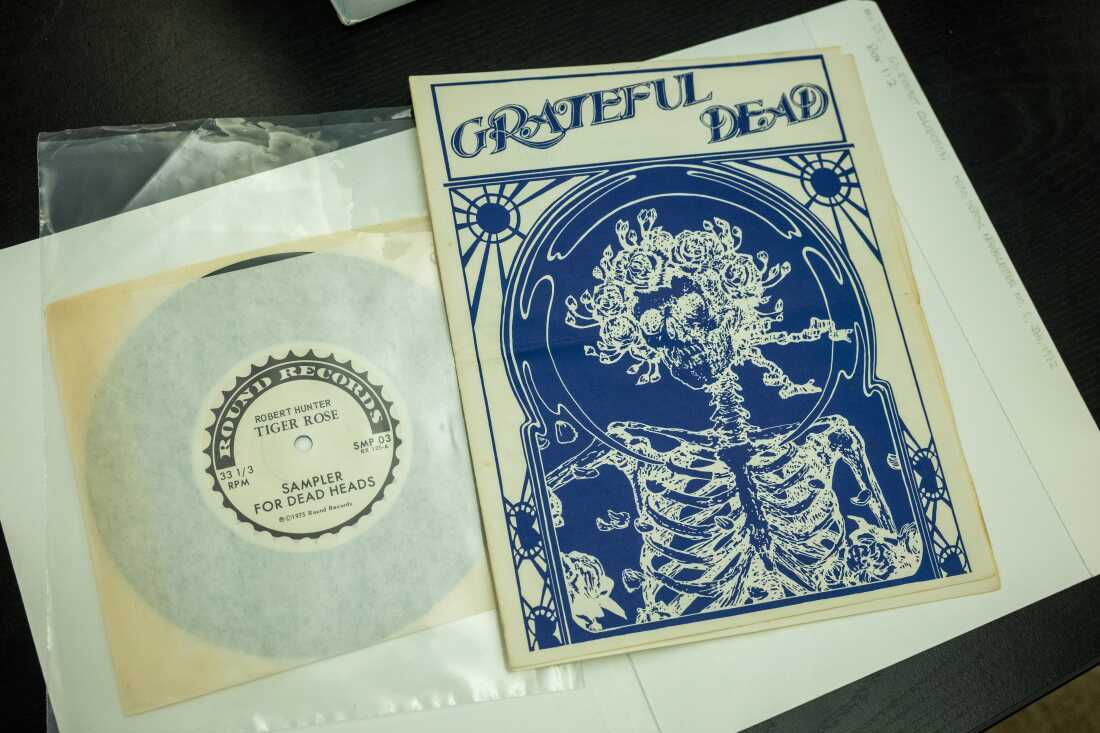 This photo shows a vinyl record in a yellowed paper sleeve. The sleeve is labeled with the words "Robert Hunter," "Tiger Rose" and "Sampler for Dead Heads." On the right side of the frame is a document that says "Grateful Dead" at the top and features an illustration of a human skeleton.